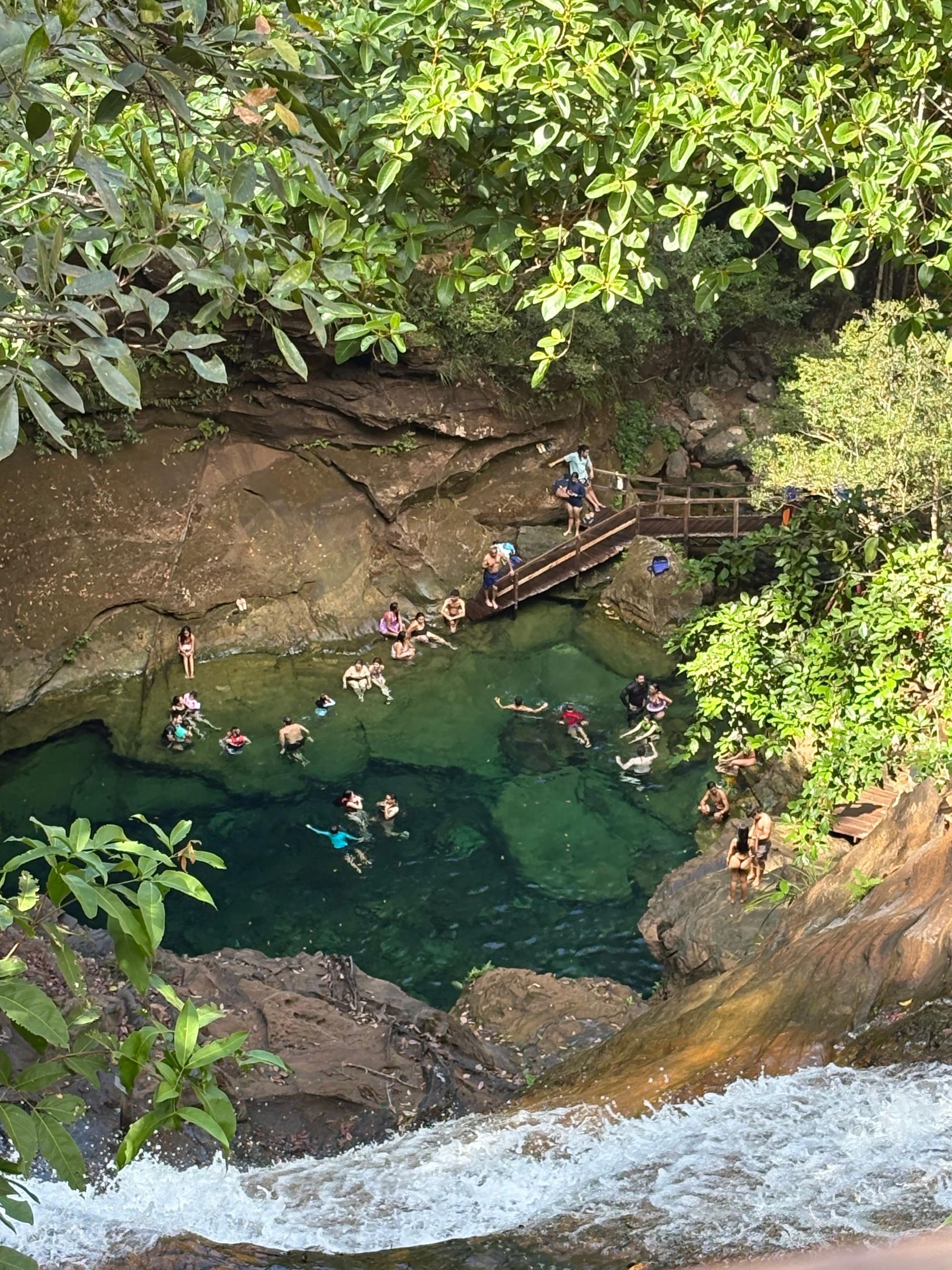 Pessoas nadando em uma piscina natural verde abaixo de uma cachoeira, cercada por rochas e árvores.