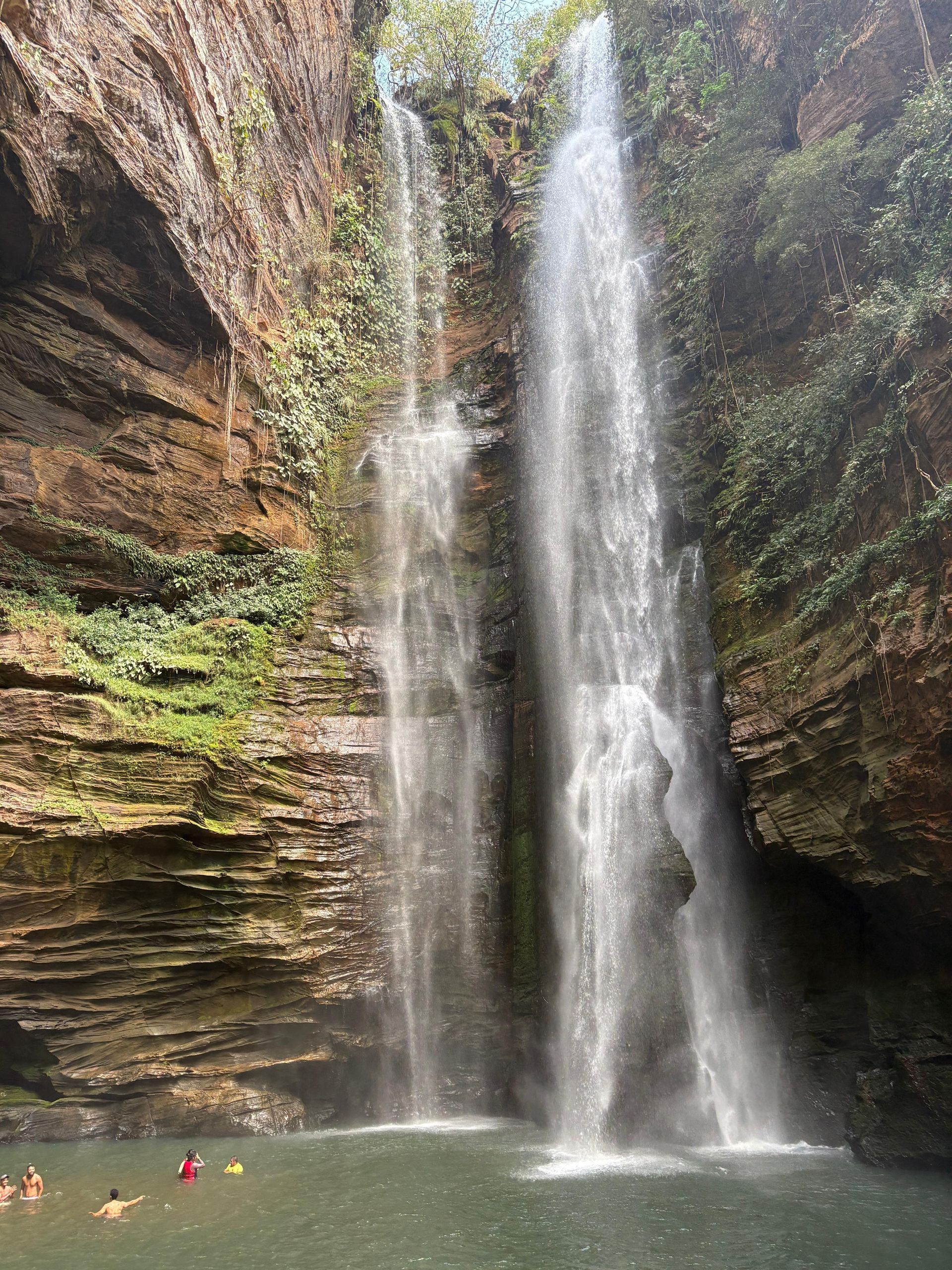 Alta cachoeira despencando em uma piscina natural em um cânion rochoso, com alguns nadadores embaixo.