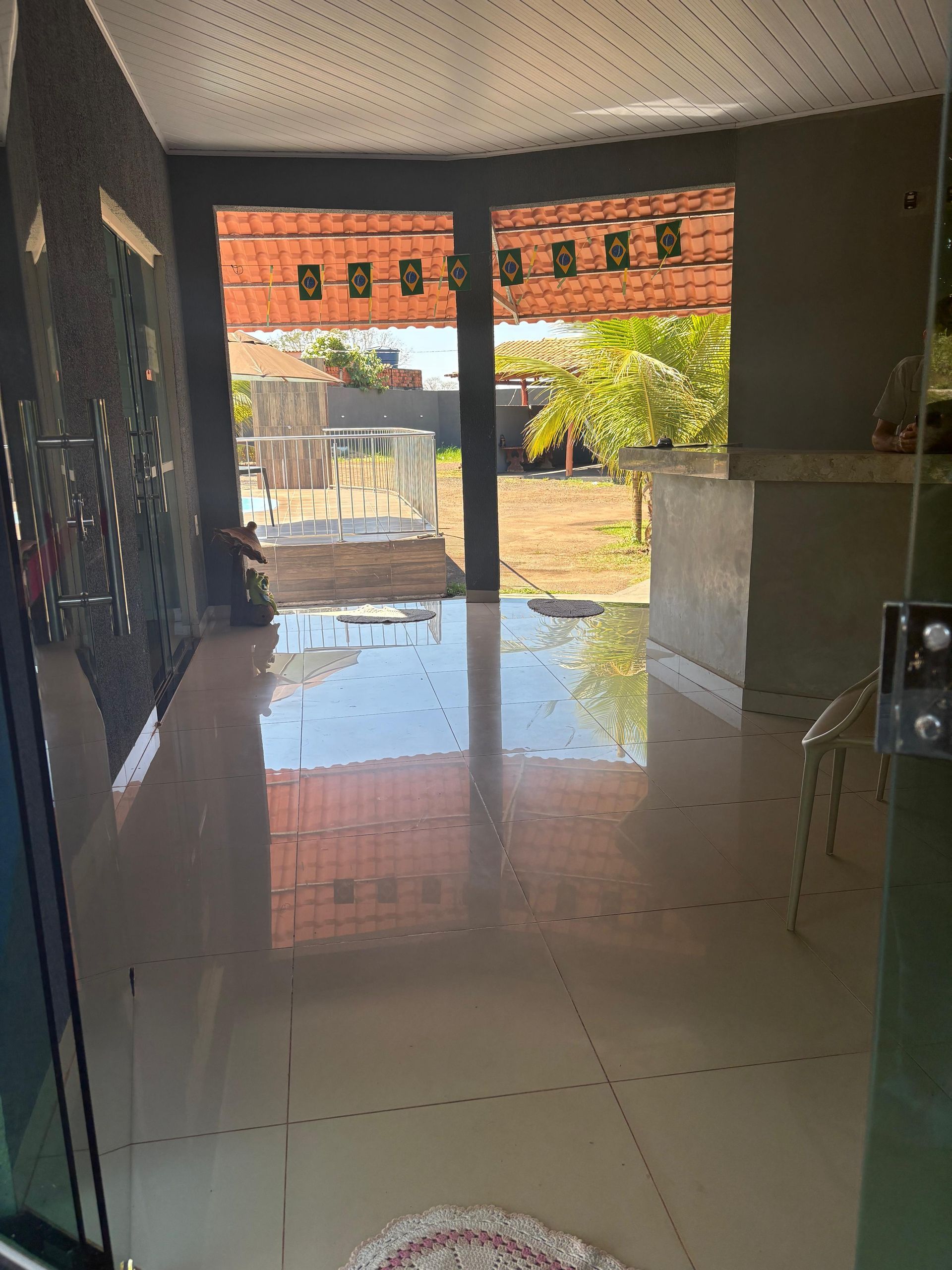 Hall de entrada com azulejos brilhantes e portas abertas, refletindo uma parede de tijolos e um pátio ensolarado do lado de fora.