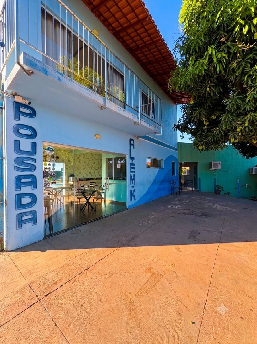 Fachada azul de uma pousada com entrada de vidro, pátio pavimentado e árvores sob luz solar intensa.