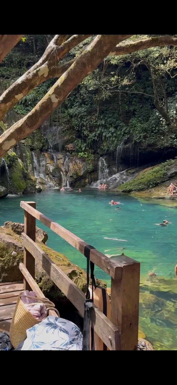 Corrimão de madeira ao lado de uma piscina natural de água turquesa, com um nadador perto de um penhasco rochoso e arborizado.