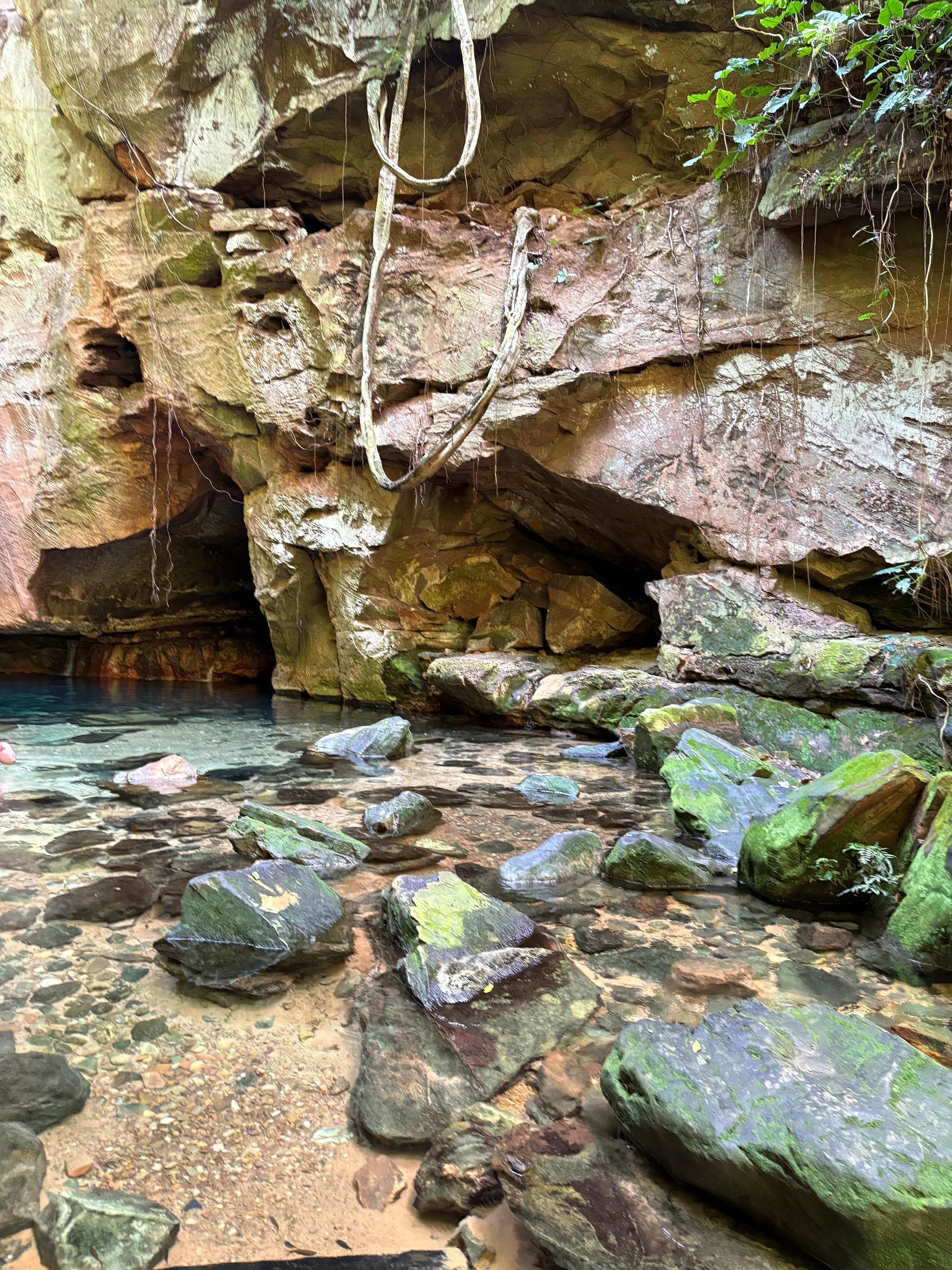 Piscina rochosa em uma caverna com água cristalina turquesa, pedras cobertas de musgo e uma pequena cachoeira.