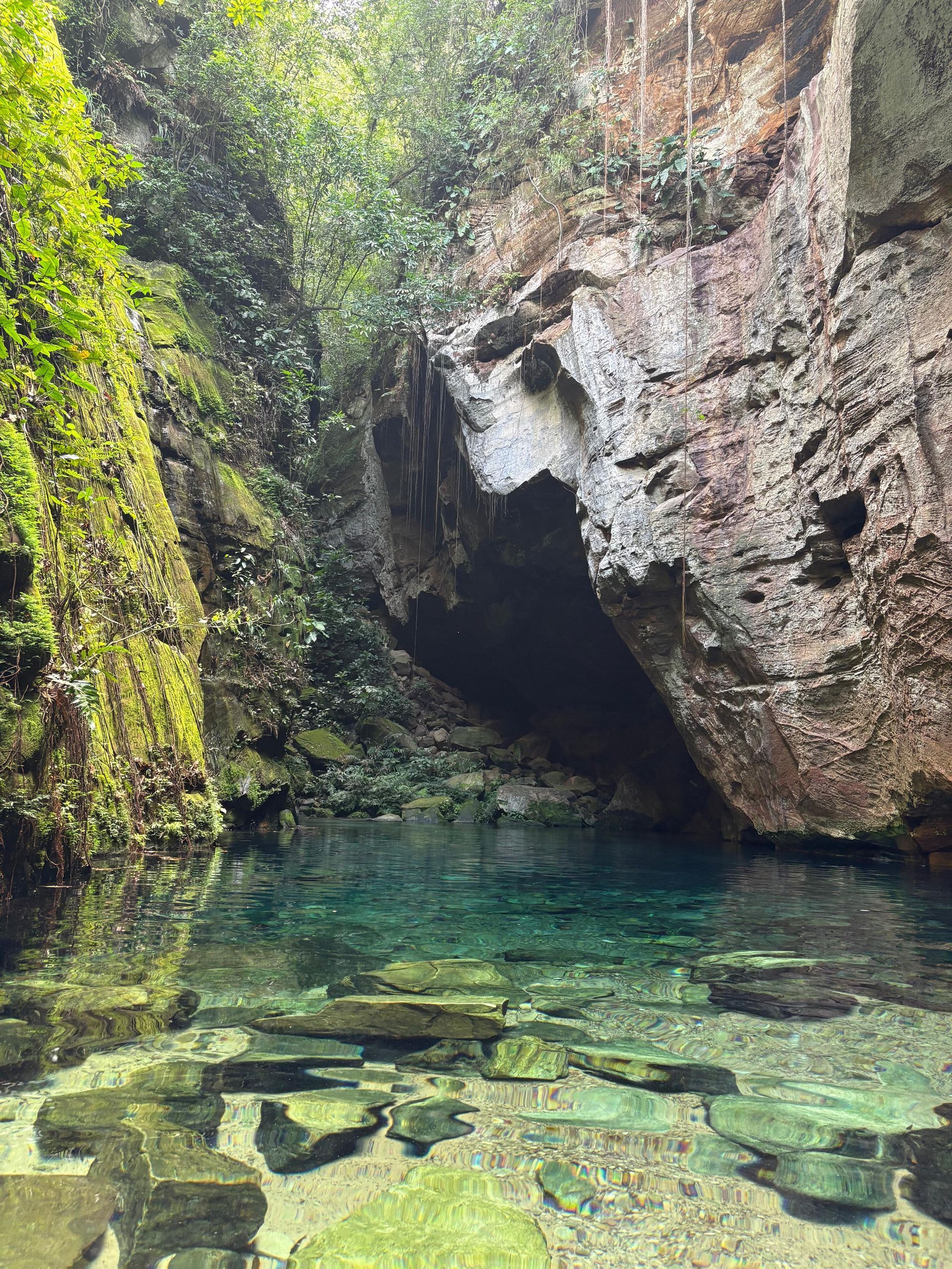Piscina cristalina de cor turquesa em uma caverna rochosa, com penhascos cobertos de musgo e a luz do sol refletida na água.