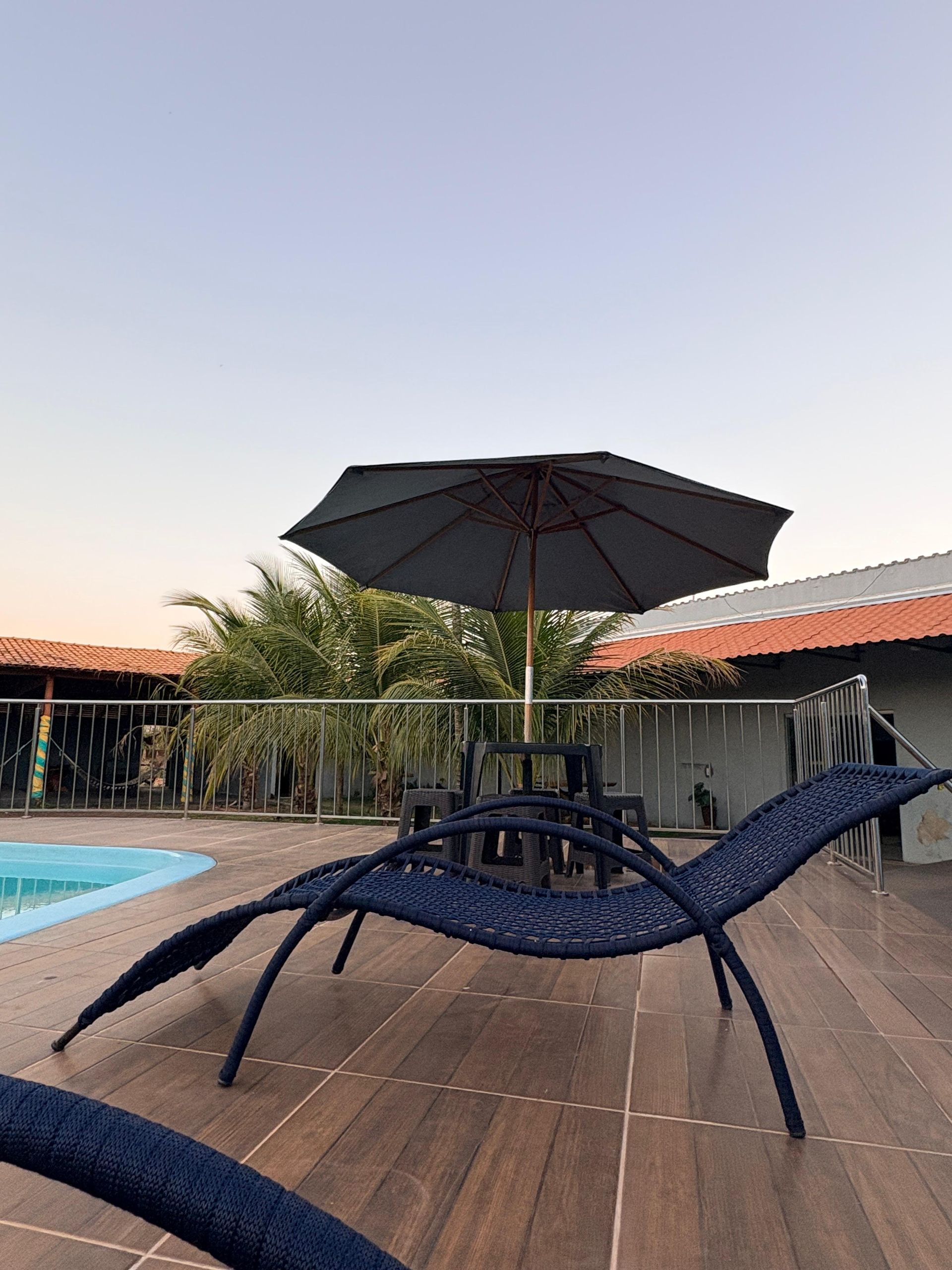 Espreguiçadeiras à beira da piscina com um guarda-sol preto em um deck de madeira ao pôr do sol.