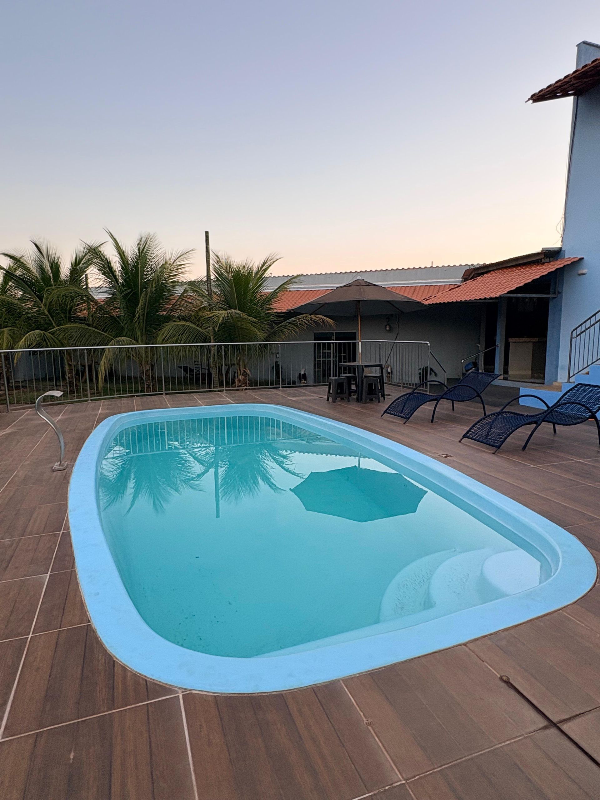 Piscina ao ar livre em um deck de madeira ao lado de uma casa ao pôr do sol, com espreguiçadeiras e palmeiras.