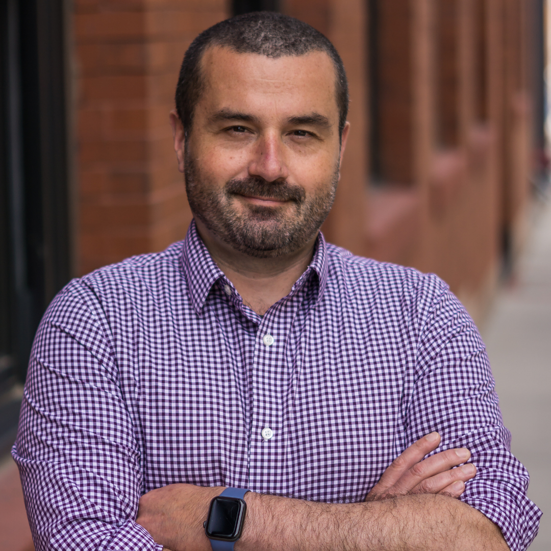 A man wearing a plaid shirt and a smart watch is standing with his arms crossed.