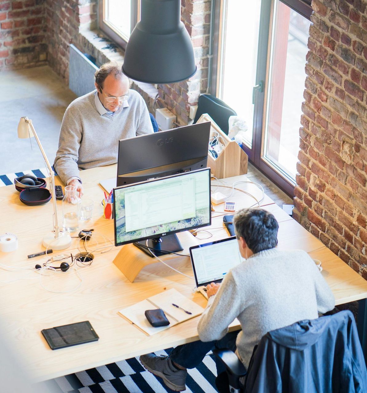 Two men are sitting at a desk with computers and laptops.