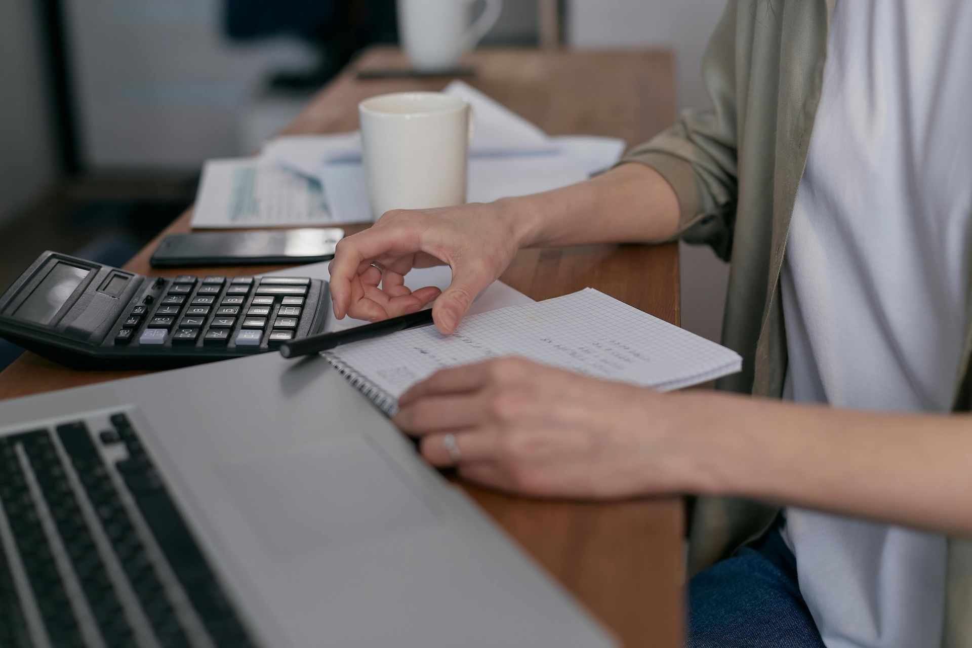 Person using calculator, laptop, and notebook at a desk. Hands hold pen near open notepad, coffee mug in background.