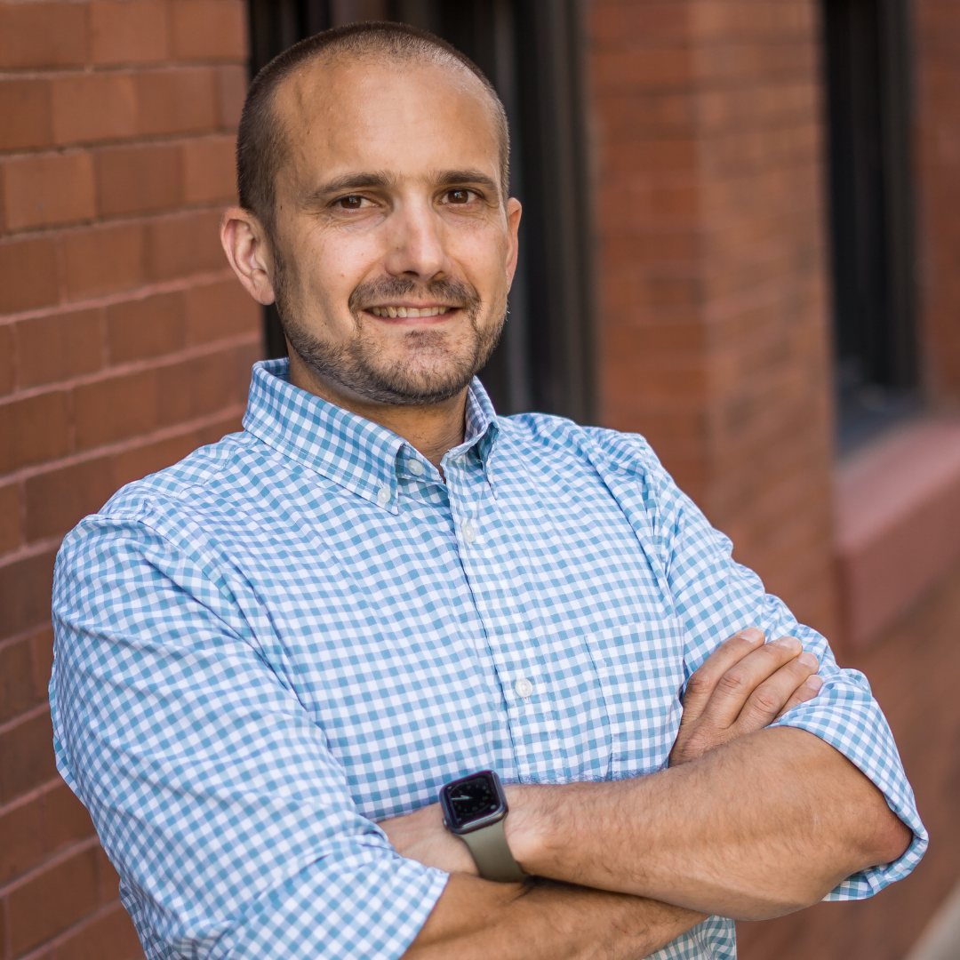A man wearing a watch is standing with his arms crossed in front of a brick wall.