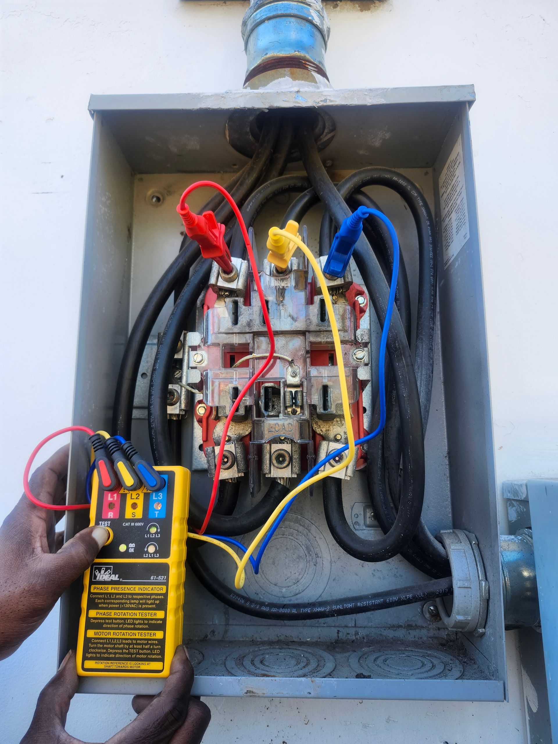 Hand holding a multimeter testing exposed electrical wiring in a gray utility box.