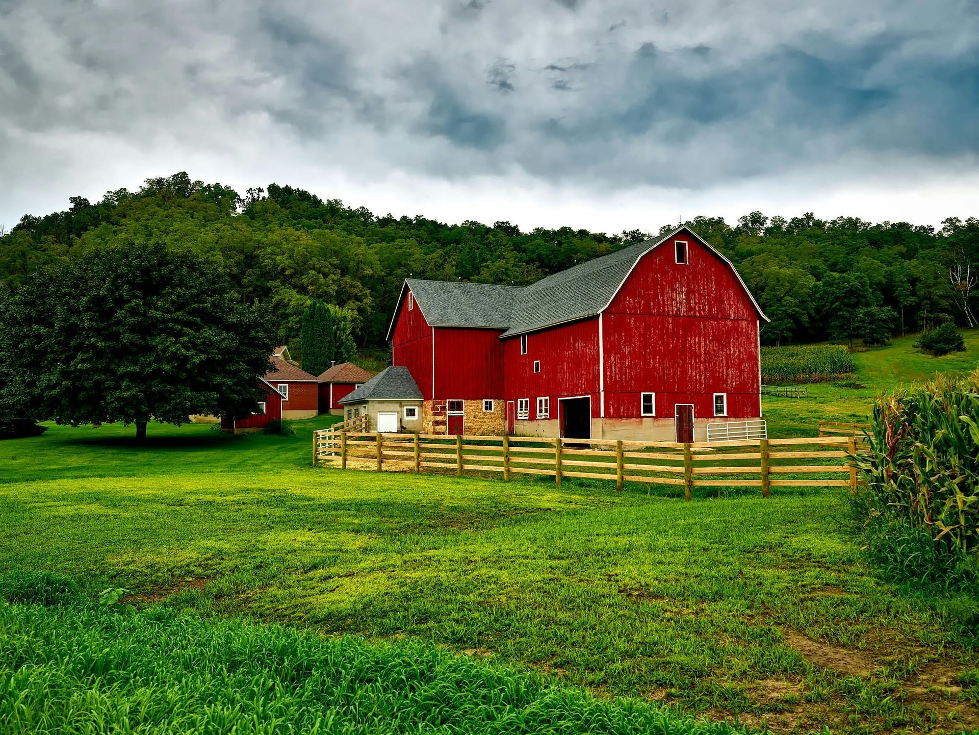 Red barn with gray roof on a green field, wooden fence. Dark clouds overhead, trees in the background.