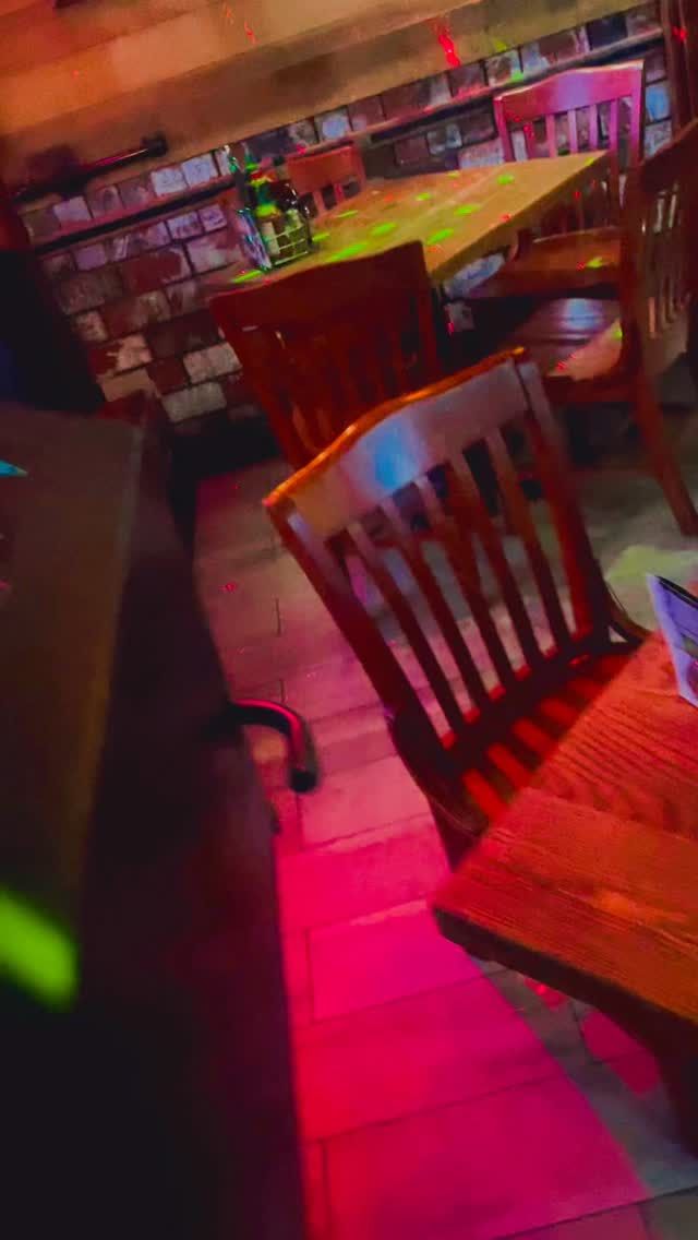 Interior of a warmly lit restaurant. Wooden tables and chairs, colorful neon lights reflecting, books on shelves.