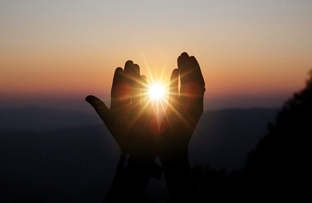 Hands cupped around the sun at sunset, silhouetted against an orange and purple sky.