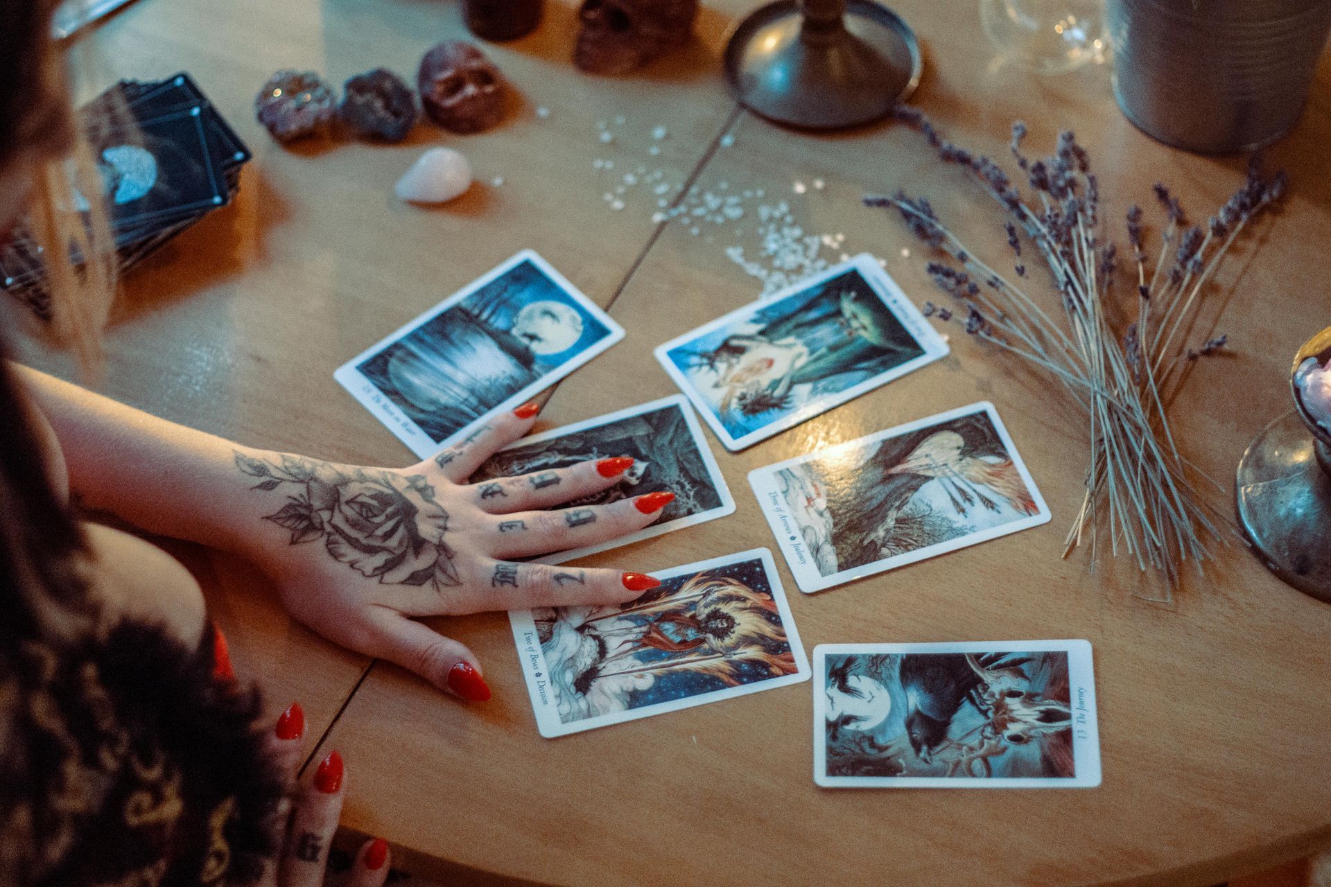 Woman's hand with tattoo and red nails laying tarot cards on a wooden table with crystals and lavender.