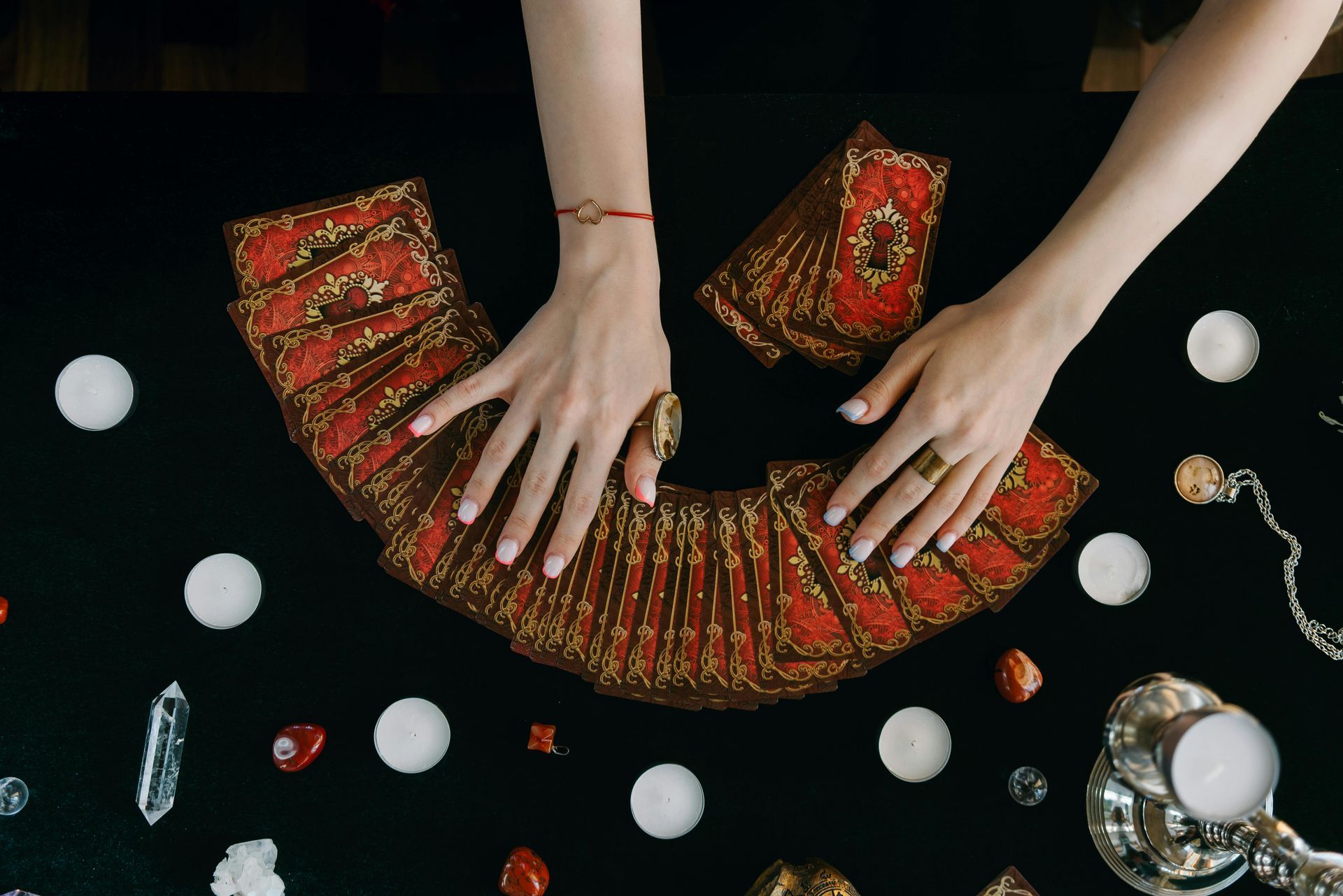 Hands over tarot cards fanned on a black surface, surrounded by candles, crystals, and jewelry.