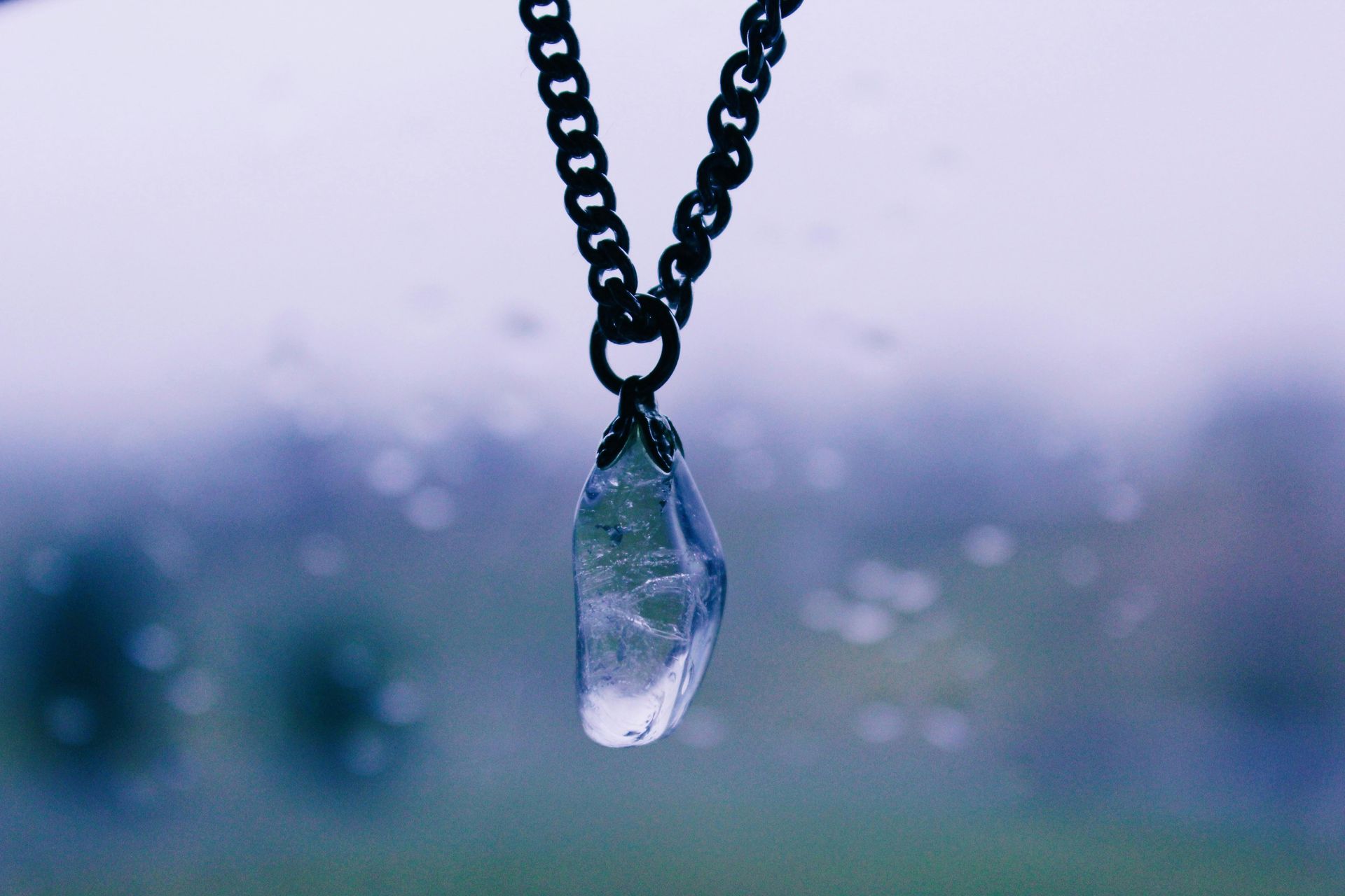 Clear crystal pendant on a black chain, hanging in front of a blurry, misty background.