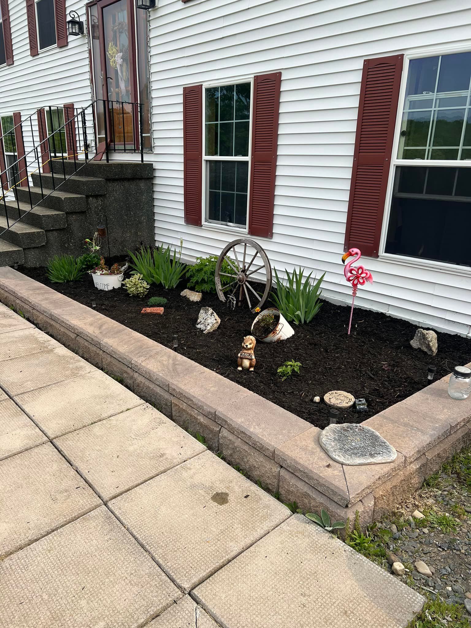 A flower bed with brown mulch, rocks, and a decorative wagon wheel in front of a white house with red shutters.