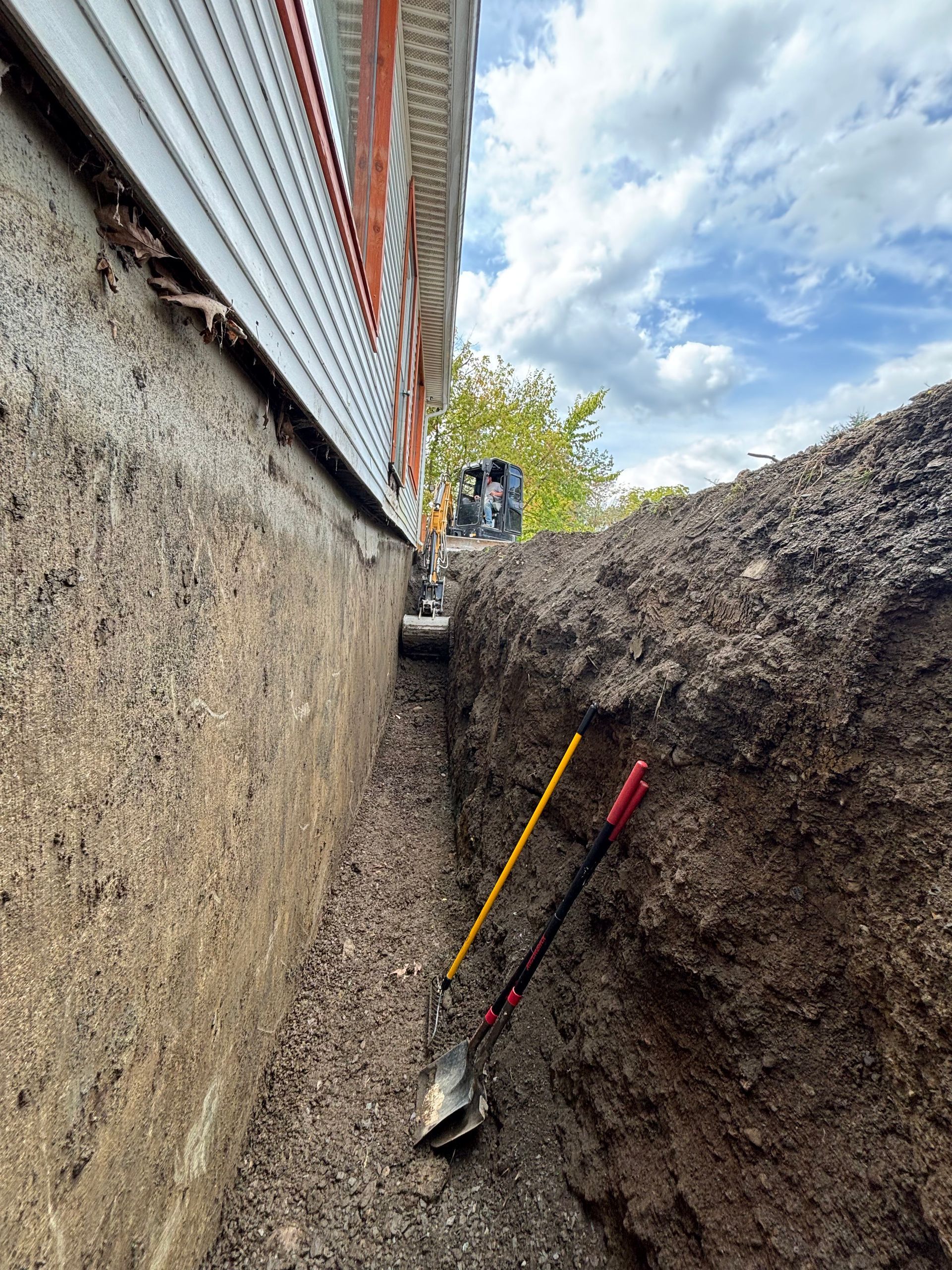 A narrow trench dug alongside a building. Shovels rest in the trench. Overcast sky.