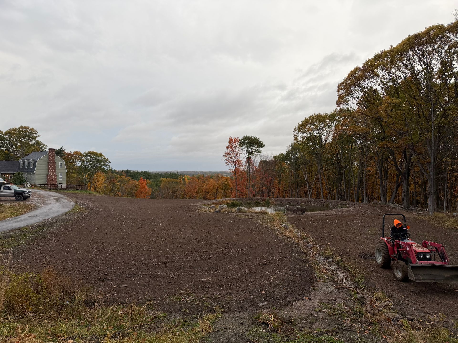 A tractor operator plowing a field near a house and trees under a cloudy sky.