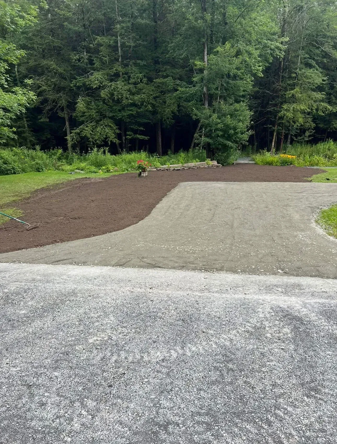 A gravel driveway transitions to a dark soil garden bed, surrounded by a forest.