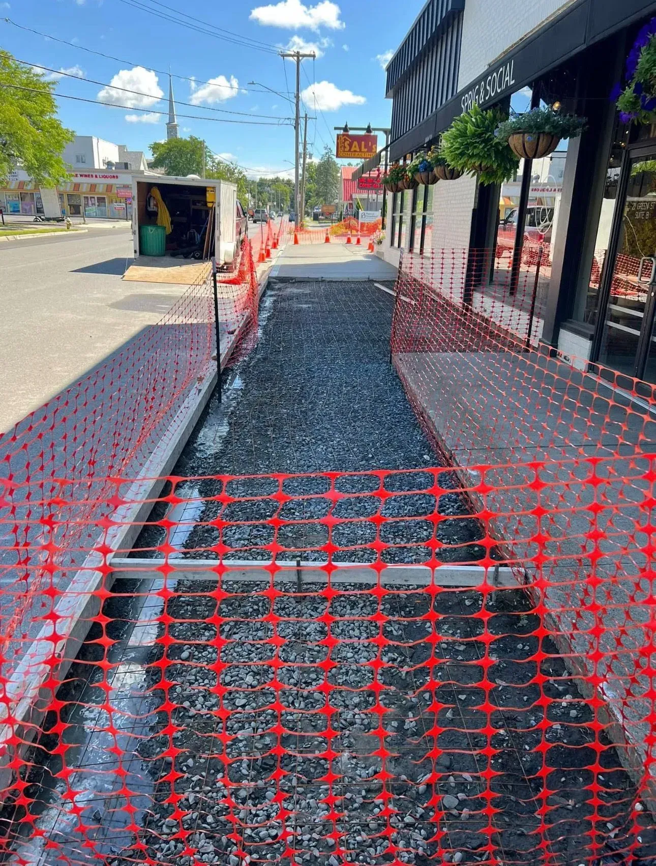 Sidewalk construction: gravel-filled trench blocked by orange safety fencing. Street and storefronts in background.
