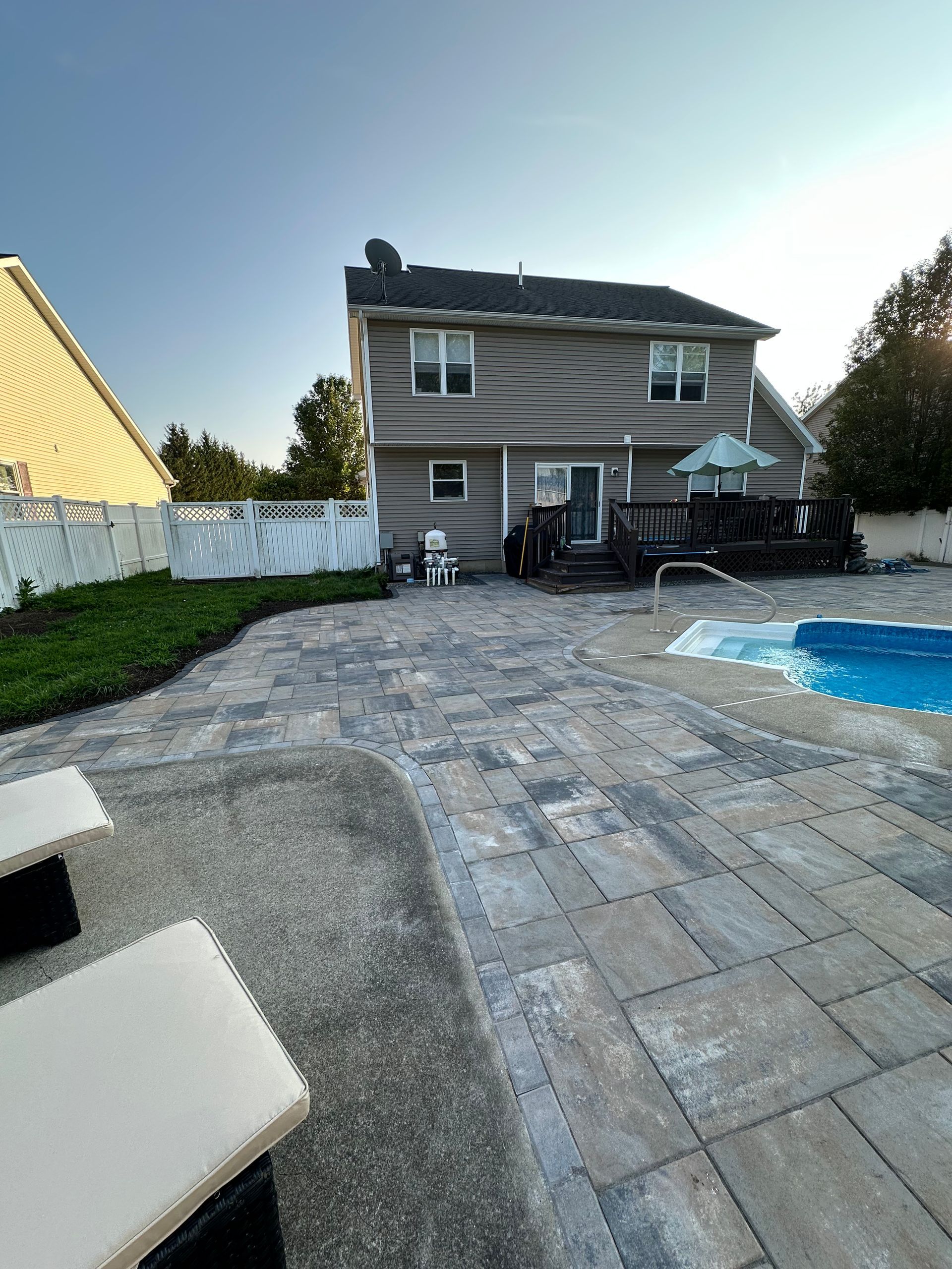 Backyard patio with pool, house, and paving stones. Blue sky.