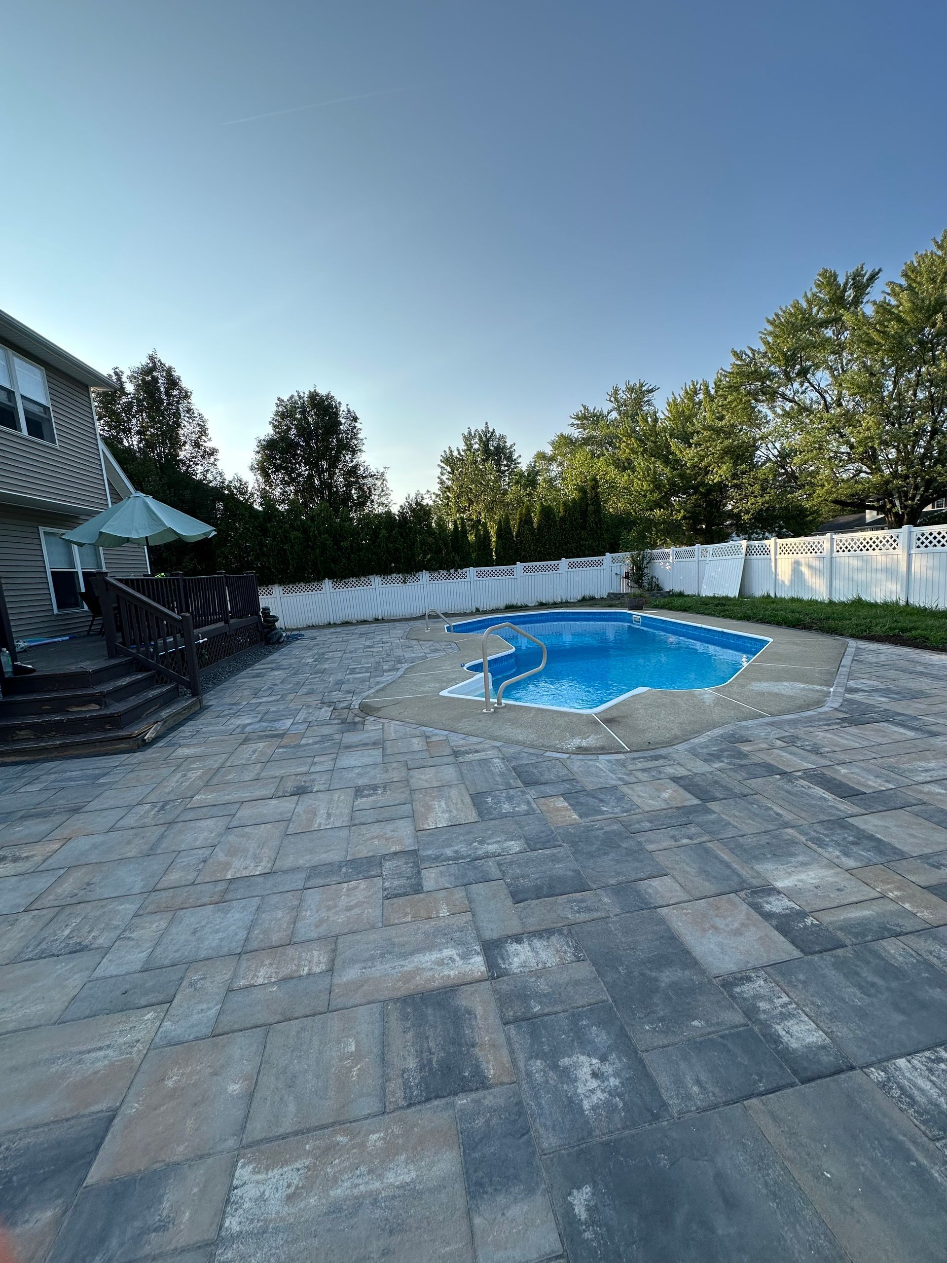 Backyard with a swimming pool and patterned stone patio; blue water, blue sky, and green trees.