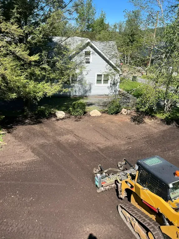 A skid steer grading dirt in front of a light gray house. Trees surround the yard.
