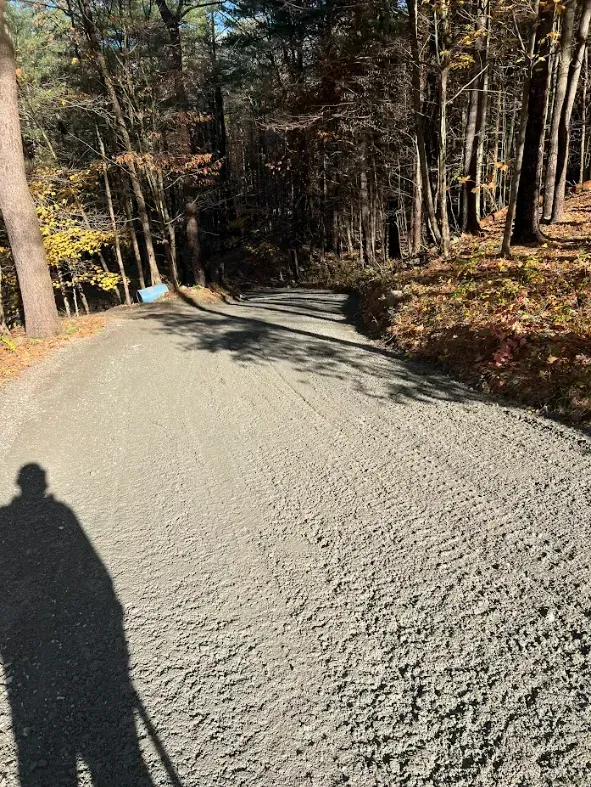 Gravel driveway curves uphill through a wooded area with trees and fall foliage. Shadow of person on left.