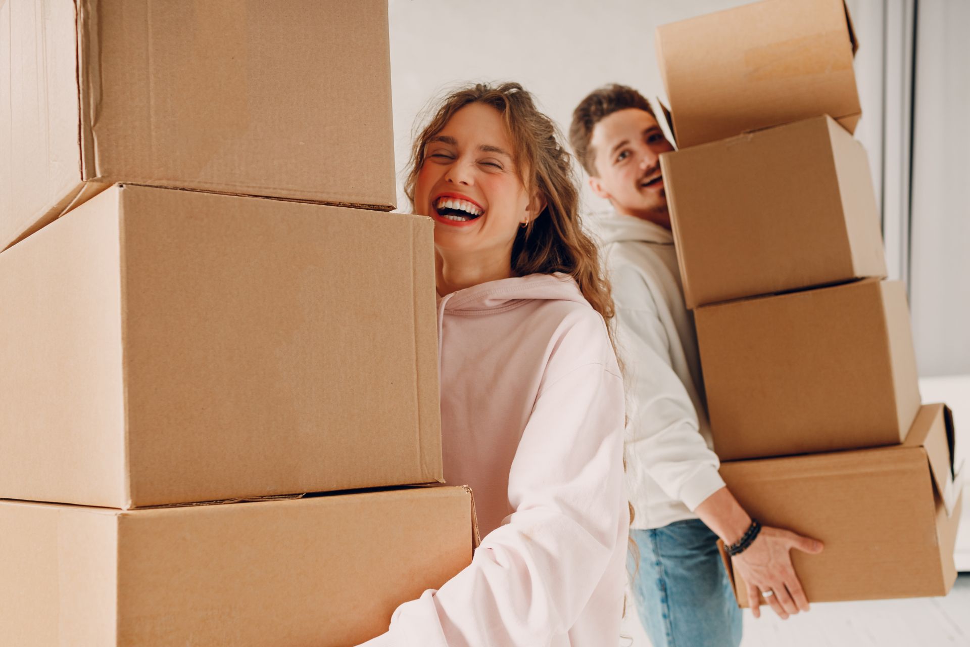 Two people smiling while carrying stacked cardboard moving boxes indoors