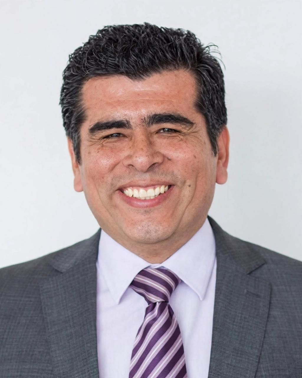 Hombre sonriente con traje gris, camisa lavanda y corbata morada a rayas, sobre un fondo claro