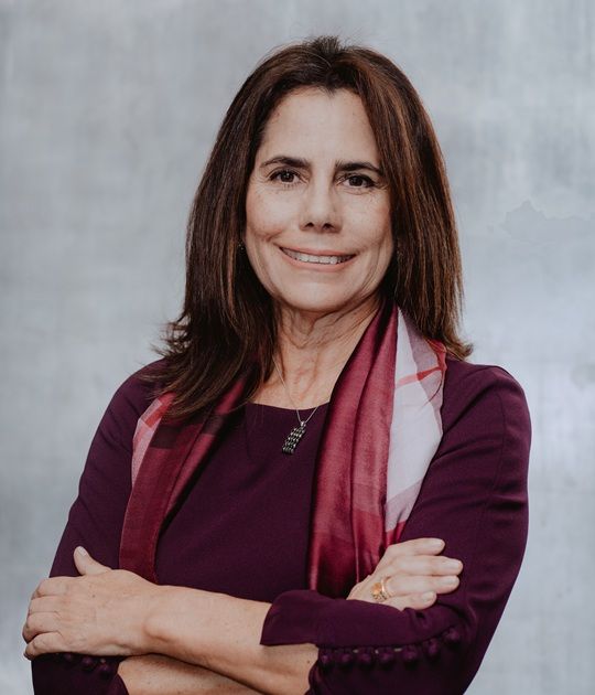 Retrato de una mujer sonriente con una blusa color burdeos y una bufanda, con los brazos cruzados frente a un fondo gris