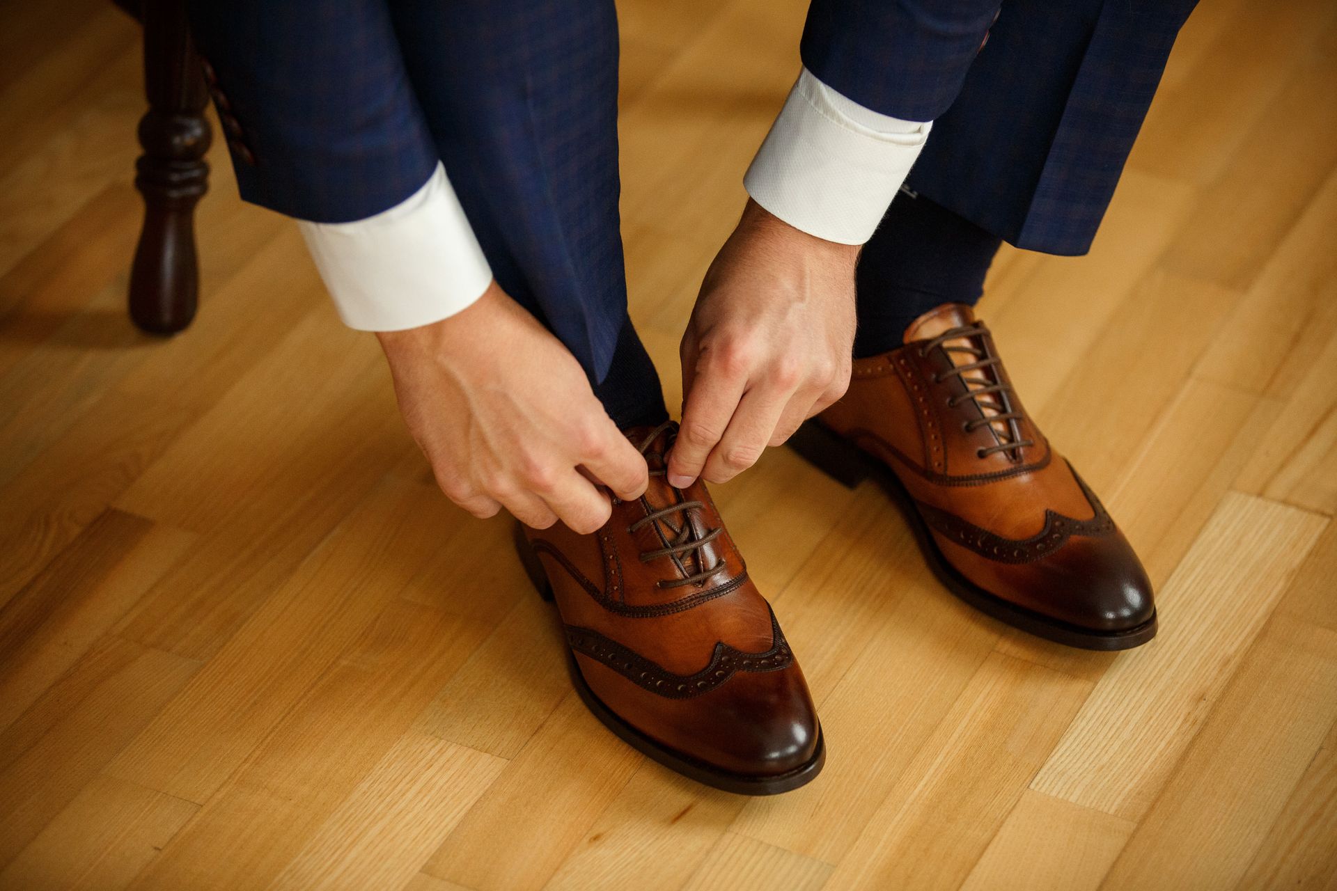 Man in suit tying brown leather dress shoes on a wooden floor.