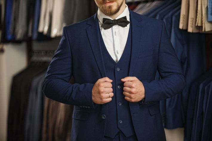 Man in a navy blue suit and bow tie, indoors in a suit shop, posing with hands on lapels.