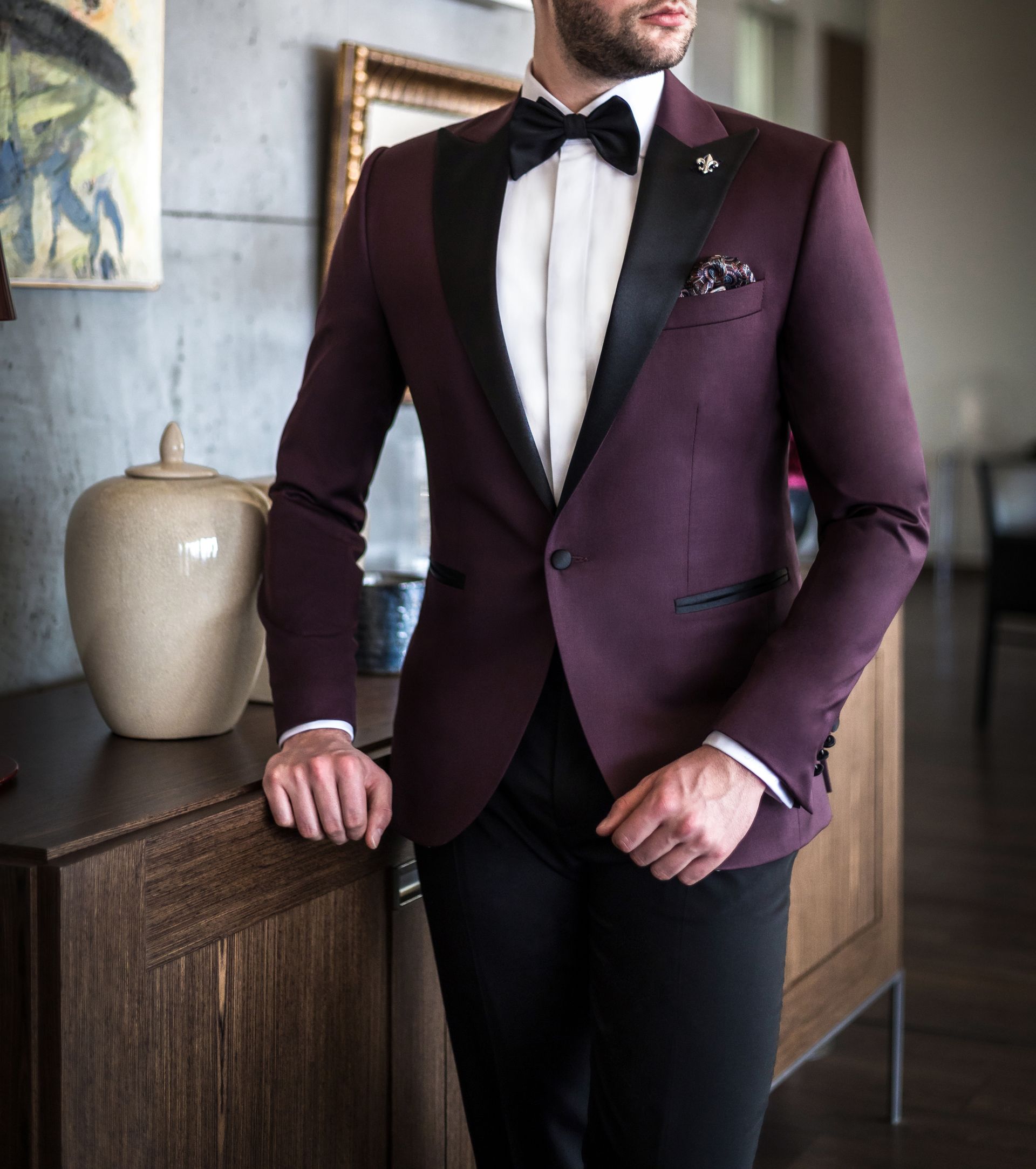 Man in a dark purple tuxedo with black lapels and bow tie, standing next to a wooden cabinet.