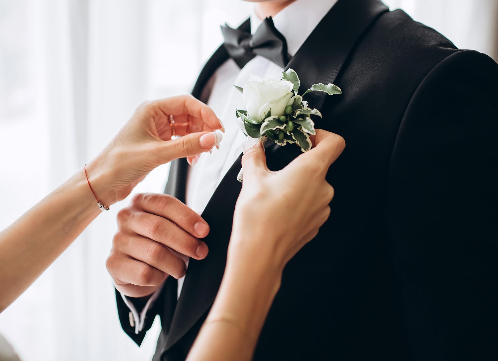 Hands adjusting a white rose boutonniere on a man's black tuxedo jacket.