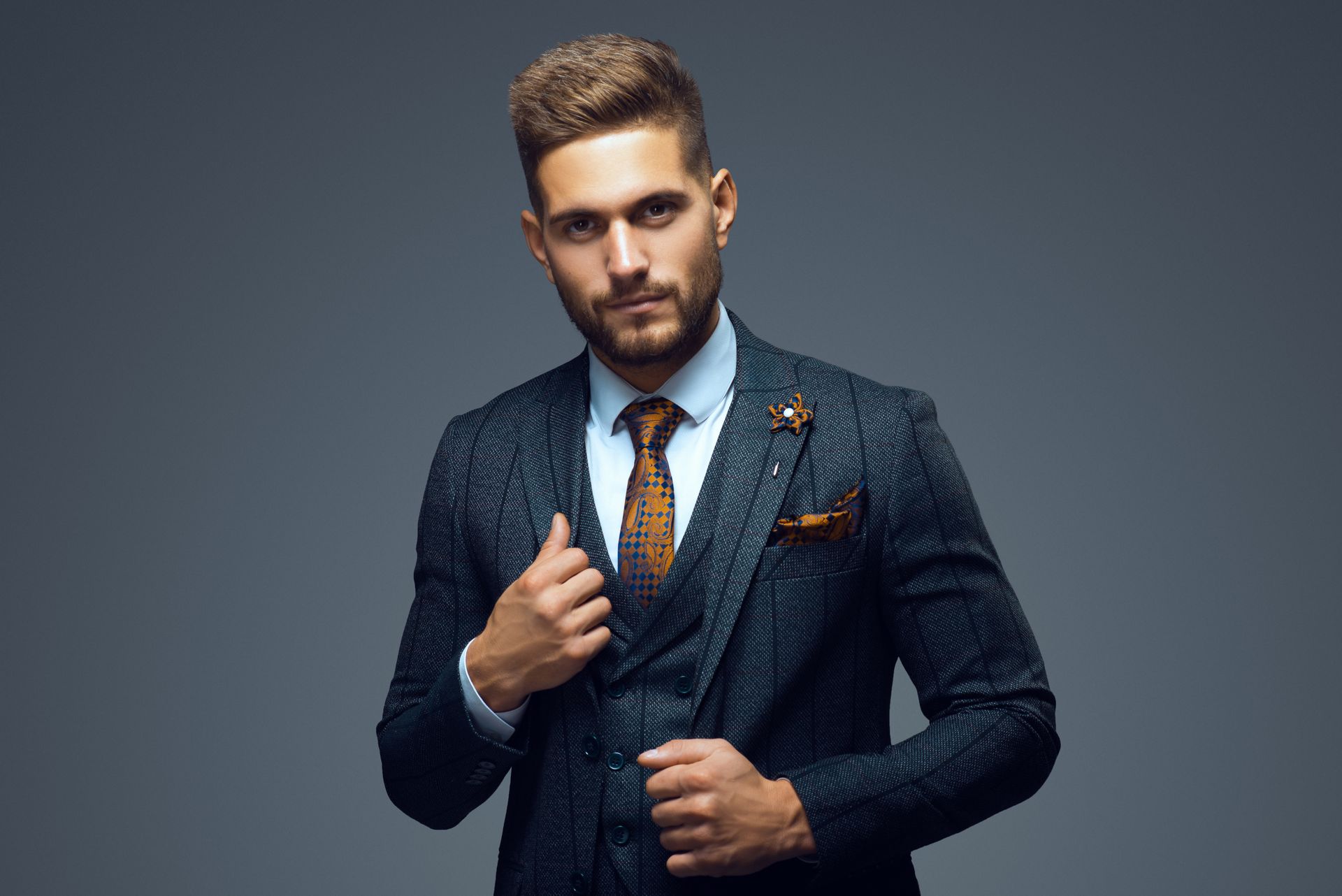 Man in a blue patterned suit and tie, adjusting his jacket; studio shot against gray background.