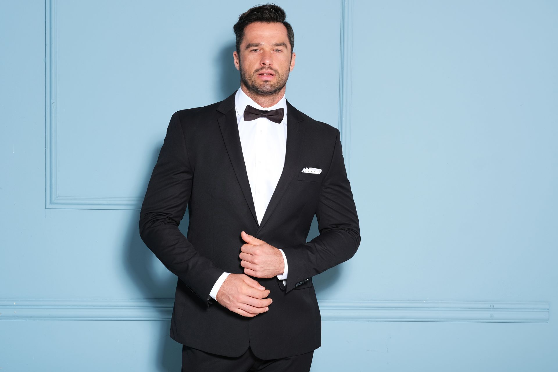 Man in black tuxedo, bow tie, white shirt, closed eyes, hands near chest, light blue wall.