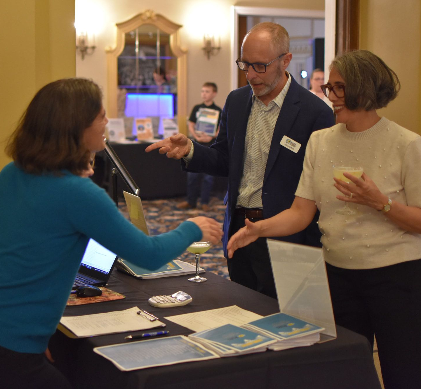 Three people at a table, two shaking hands. A woman at the table greets a man and woman. Indoors, documents and laptop visible.