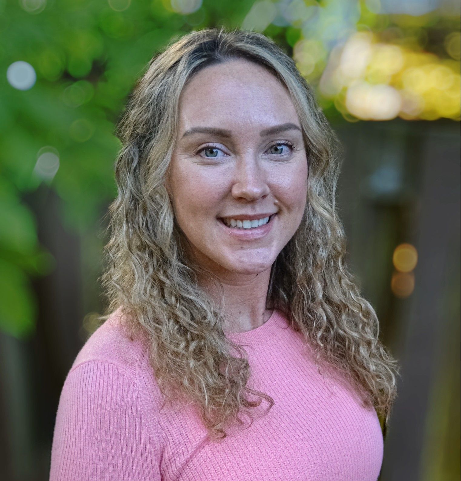 Woman with long curly blonde hair smiles wearing a pink shirt outdoors.