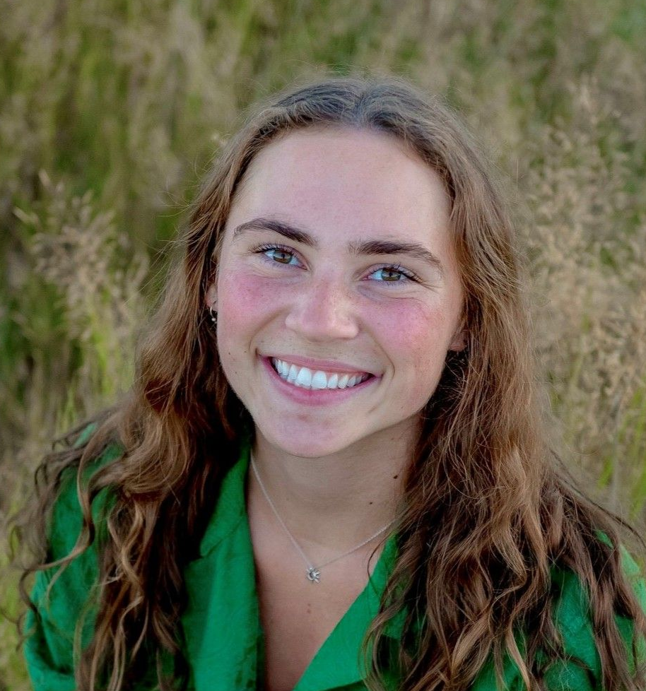 Woman with long wavy brown hair smiles, wearing a green jacket, in a field of tall grass.