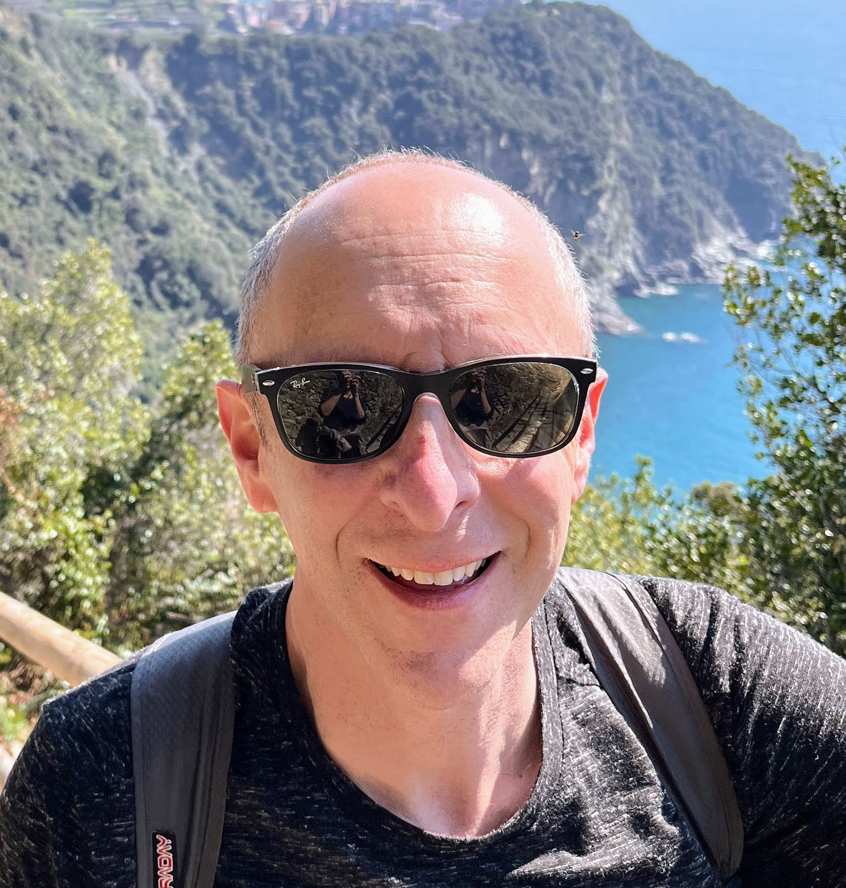 Bald man with sunglasses smiles at the camera, backpack on, backdrop of ocean and mountains.