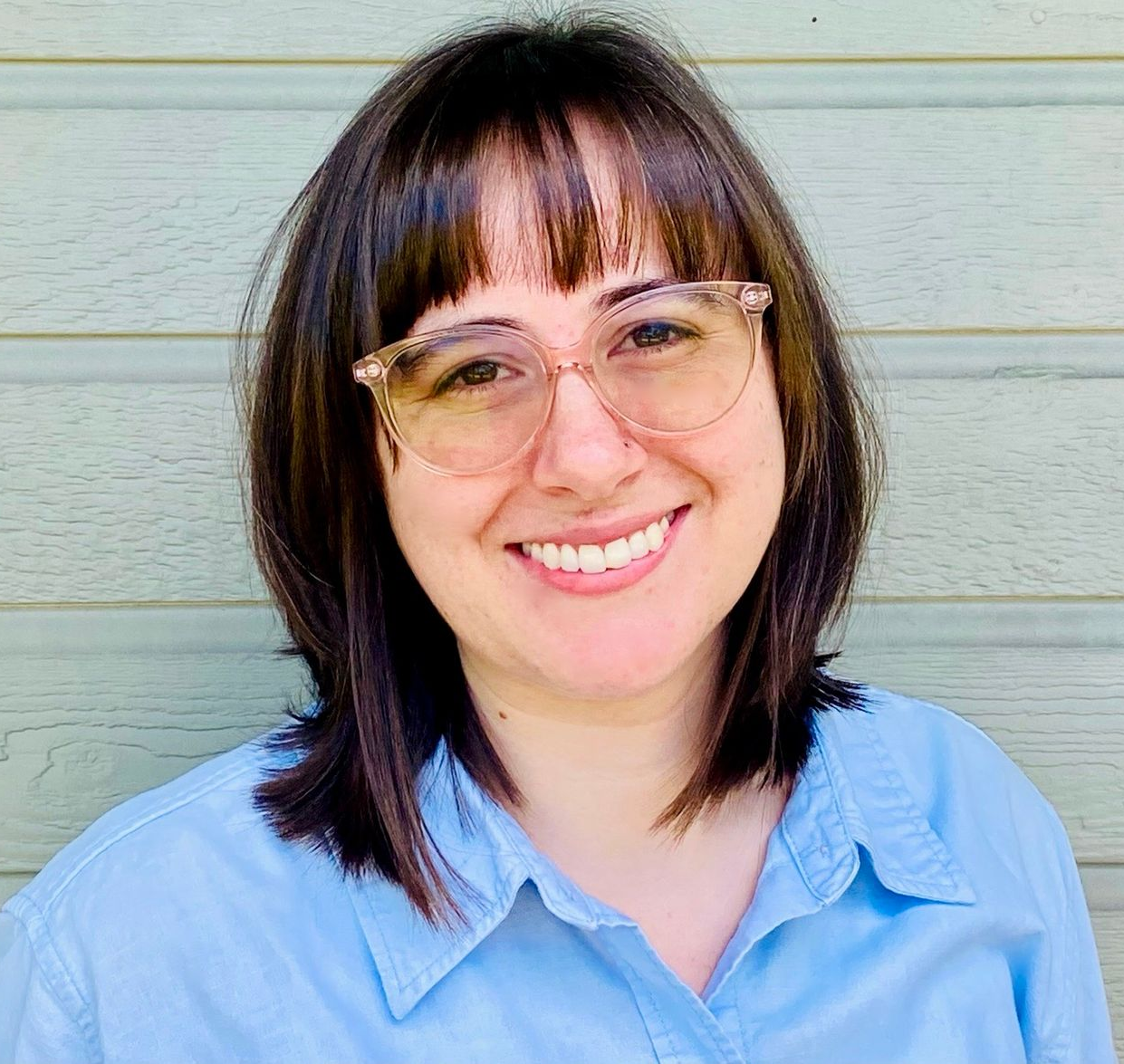 Woman with brown hair and glasses smiling, wearing a blue shirt, in front of a green backdrop.