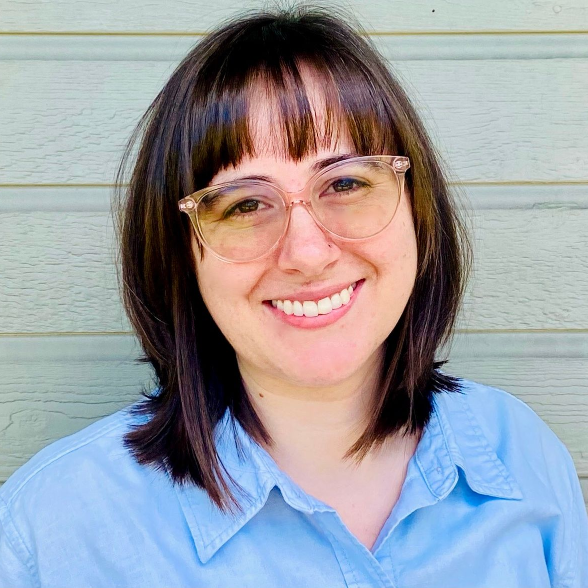 Woman with brown hair and glasses smiling, wearing a blue shirt, in front of a green backdrop.