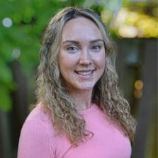 Woman with long curly blonde hair smiles wearing a pink shirt outdoors.