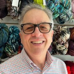 Man with glasses smiling in front of yarn shelves.