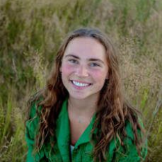 Woman with long wavy brown hair smiles, wearing a green jacket, in a field of tall grass.