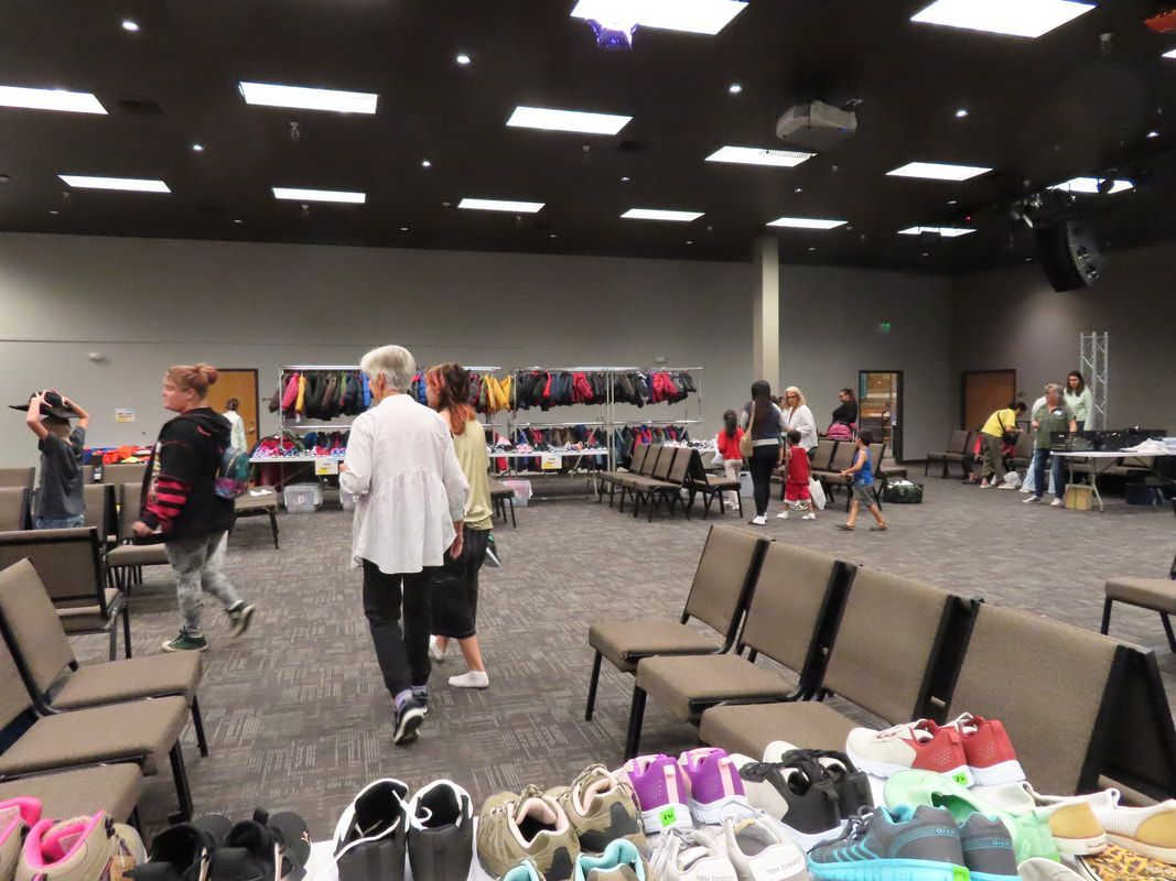 People selecting shoes in a large room. Shoes on tables. People walking around, some near racks of jackets.