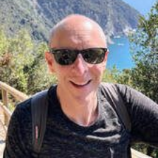 Man wearing sunglasses smiles outdoors; mountains and water in background.