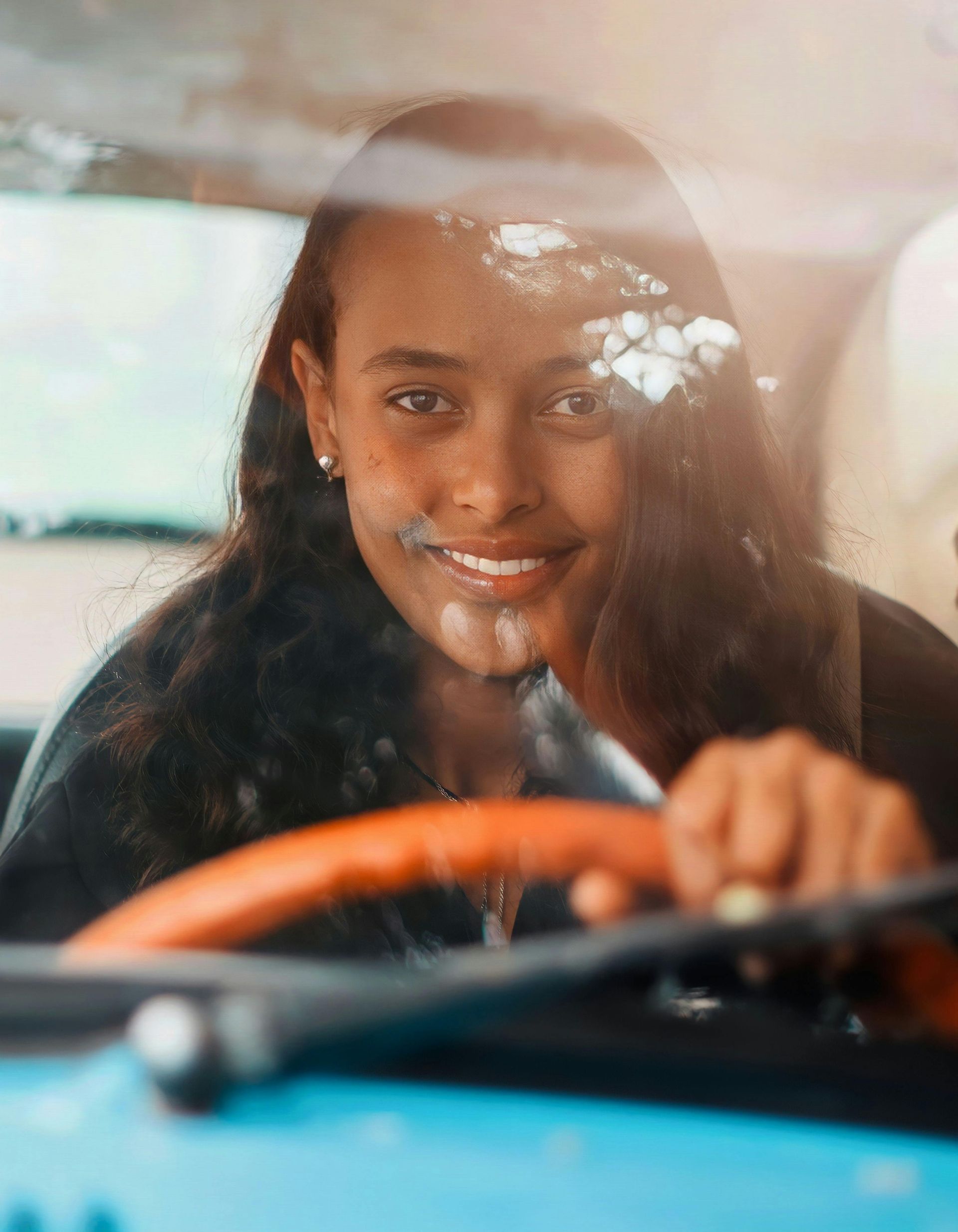 Woman smiles, holding a steering wheel, seen through a car window.